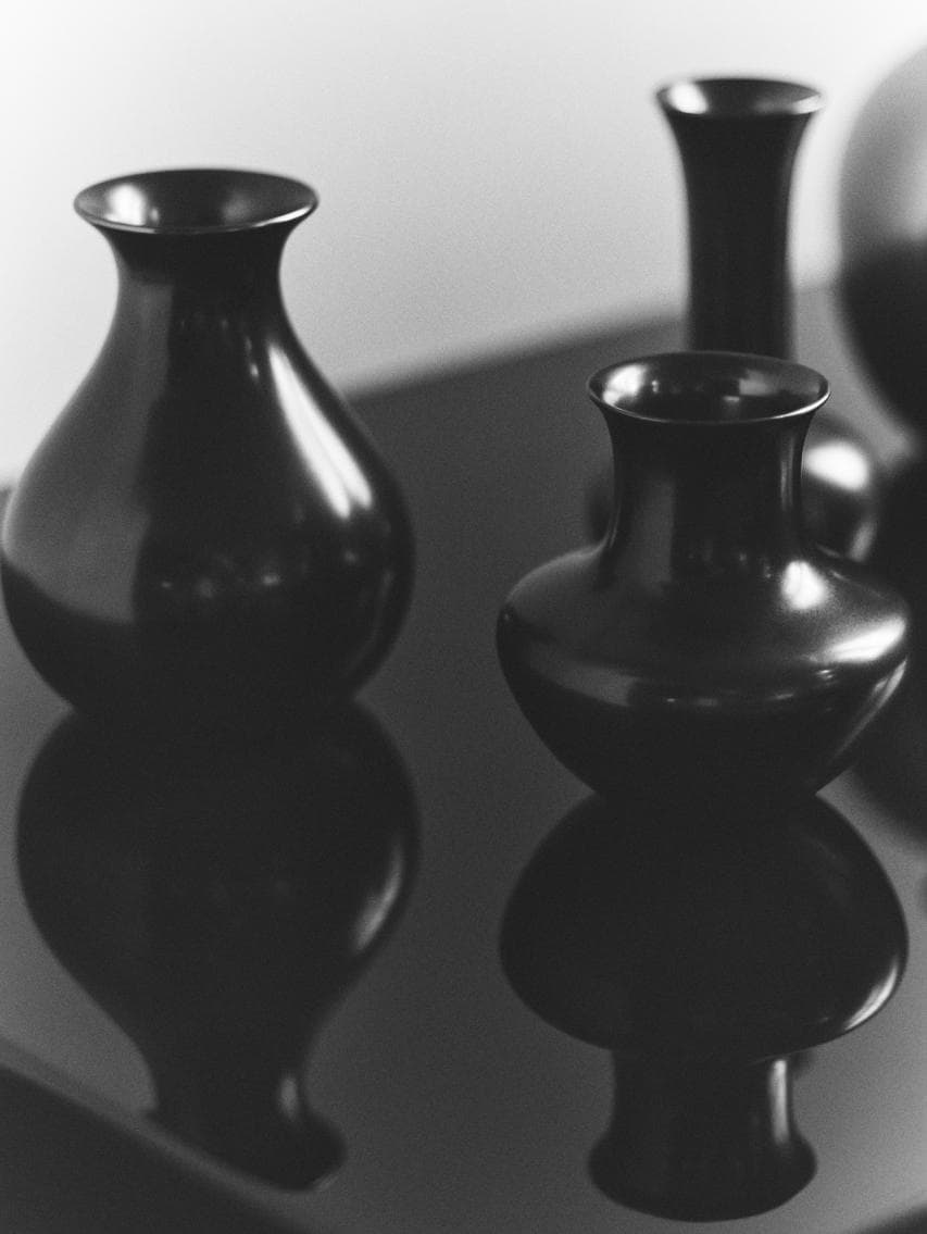 Un ensemble de vases noirs brillants aux formes arrondies et allongées, disposés sur une surface réfléchissante dans un décor monochrome minimaliste.