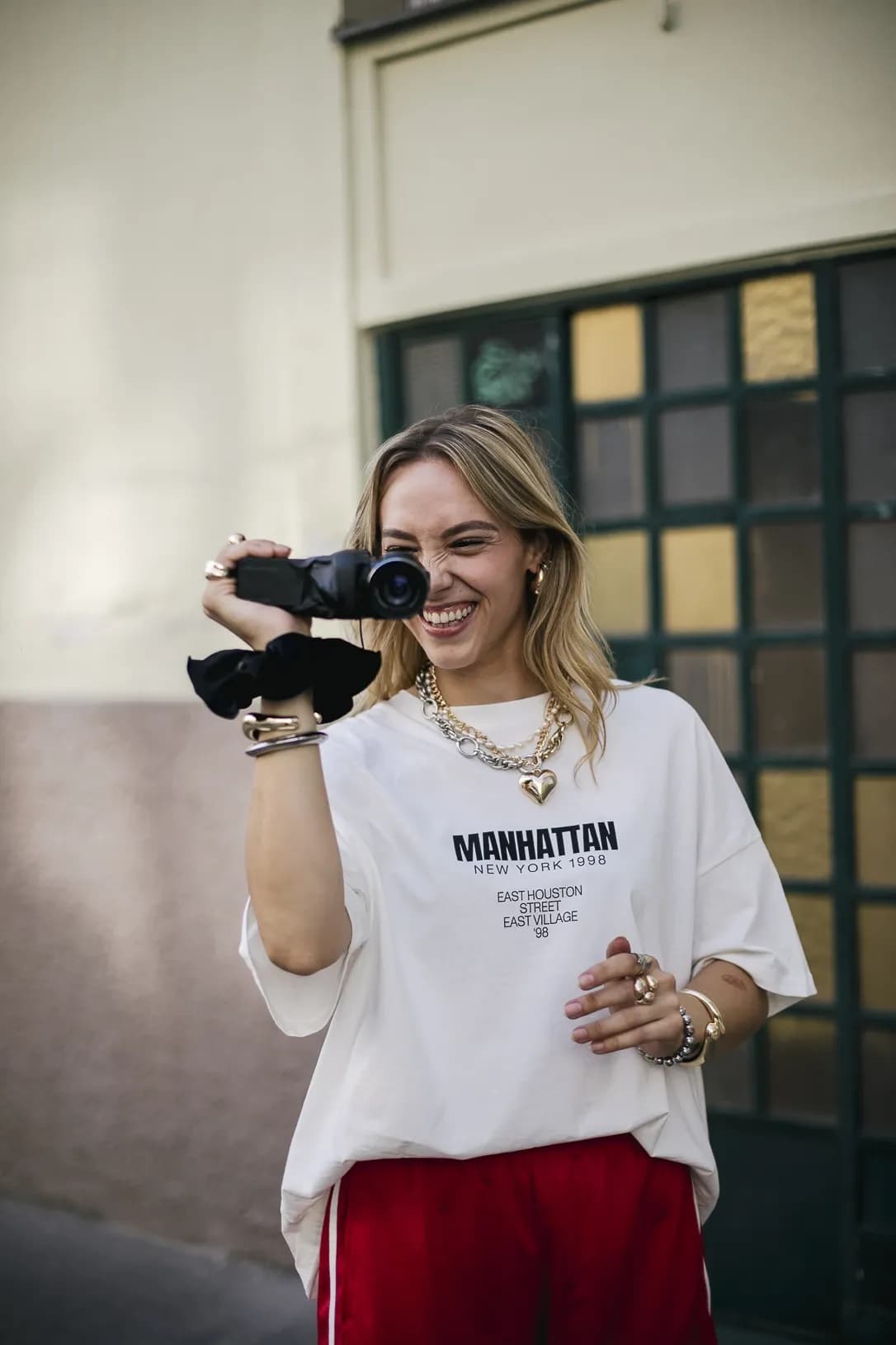 Eine lächelnde Frau in einem übergroßen T-Shirt mit Aufdruck und einer auffälligen roten Hose filmt mit einer Handkamera. Ihr verspielter, urbaner Look wird durch mehrere Schmuckstücke abgerundet.
