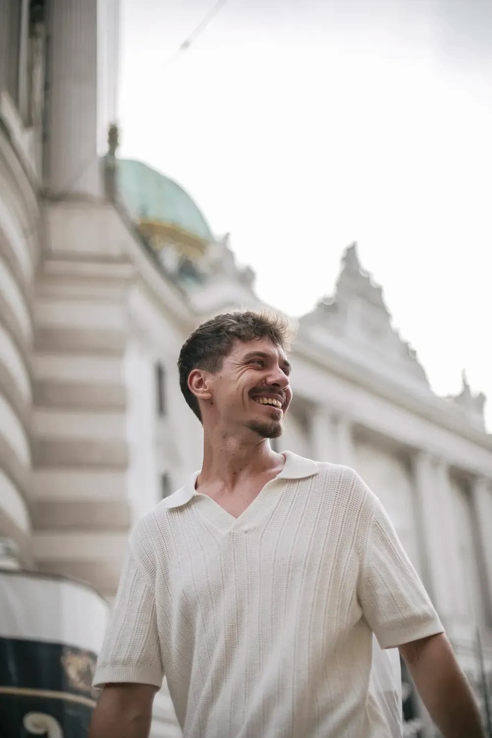 Ein lächelnder Mann in einem hellen Strickpolohemd blickt zur Seite und steht im Freien vor klassischer Stadtarchitektur.