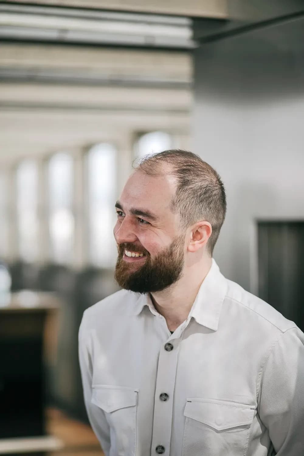 Ein lächelnder, bärtiger Mann in einem hellen Hemd mit Knöpfen blickt in einem hellen Innenraum zur Seite.