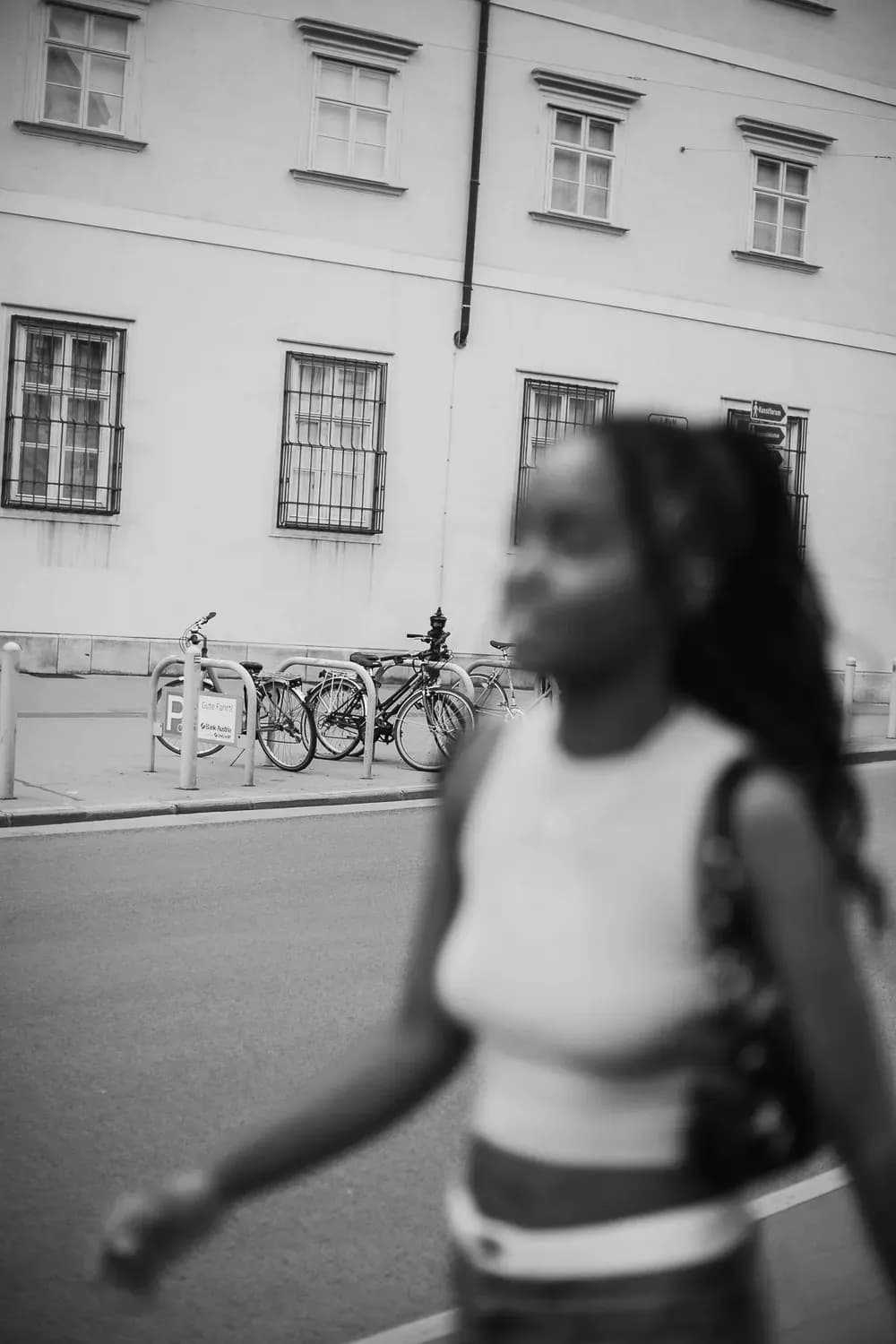Verschwommene Darstellung einer Frau, die an geparkten Fahrrädern auf einer Stadtstraße vorbeigeht; im Hintergrund ist ein klassisches Gebäude zu sehen.
