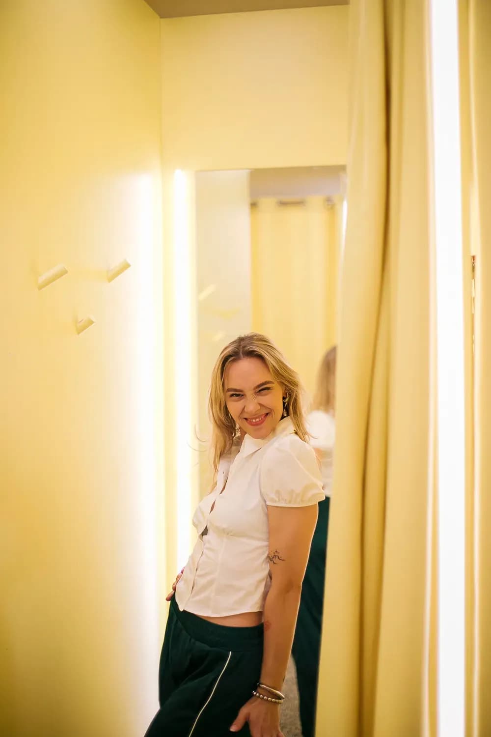 Eine lächelnde Frau in einer taillierten weißen Bluse posiert in einer sanft beleuchteten Umkleidekabine mit Spiegeln.