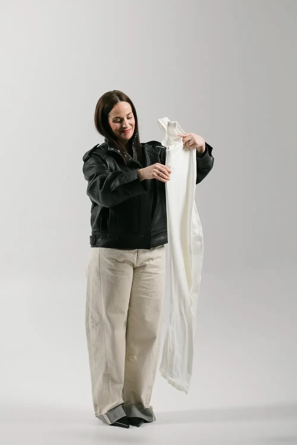 Persona con chaqueta de cuero negra sosteniendo y examinando una prenda blanca larga sobre un fondo de estudio limpio.