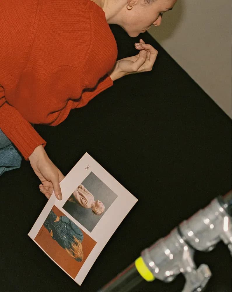 Una persona con un suéter rojo revisa imágenes de moda impresas junto a equipos de estudio en un espacio de trabajo creativo.