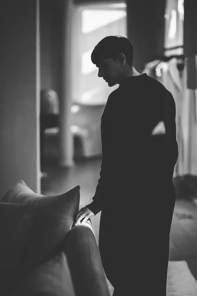 Persona de pie junto a un sofá en un interior con iluminación tenue, en una escena sombría en blanco y negro.
