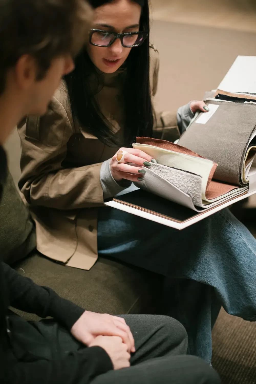Two people reviewing fabric and material samples together from an open design portfolio in a fashion studio setting.