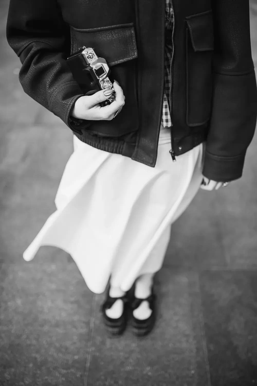 Close-up of a person wearing a dark leather jacket and flowing skirt, holding a compact camera in a monochrome fashion-style composition.