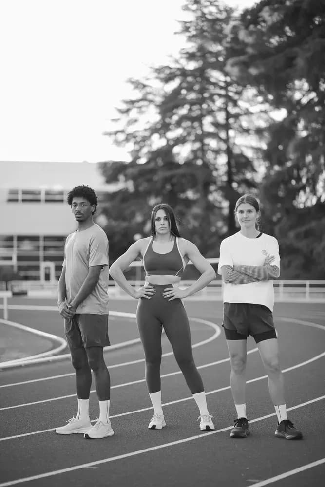 Tres atletas de pie sobre una pista de atletismo, vestidos con ropa deportiva, posan con confianza en una escena al aire libre en blanco y negro.