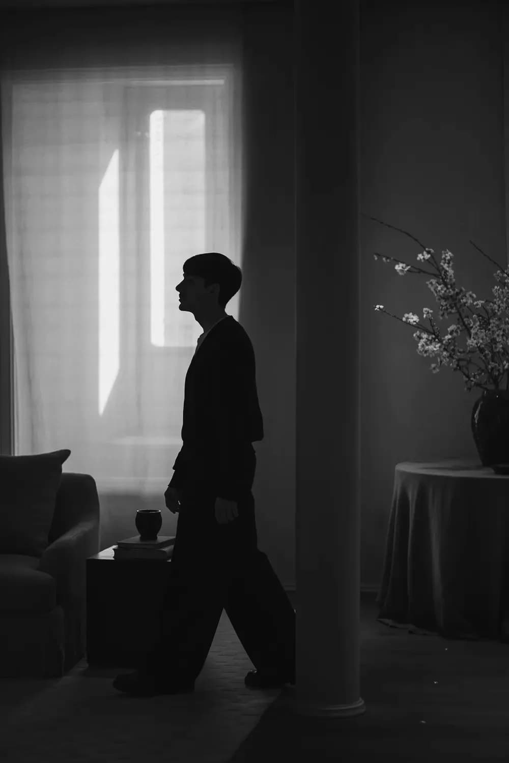 Silhouette of a person walking through a softly lit interior space with large windows, minimalist furniture, and floral decor in a monochrome setting.