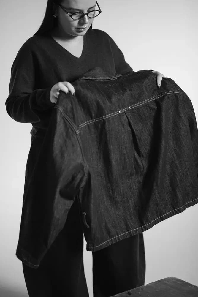 Una persona con gafas sostiene e inspecciona una chaqueta vaquera oscura en un estudio minimalista en blanco y negro.