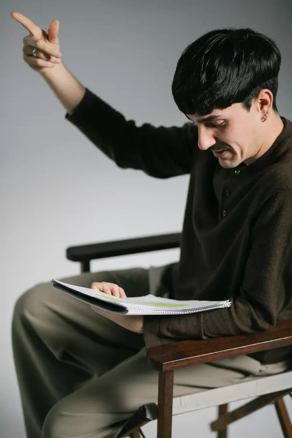 Person sitting in a chair reading from a notebook while gesturing upward, wearing dark casual clothing in a minimalist studio setting.