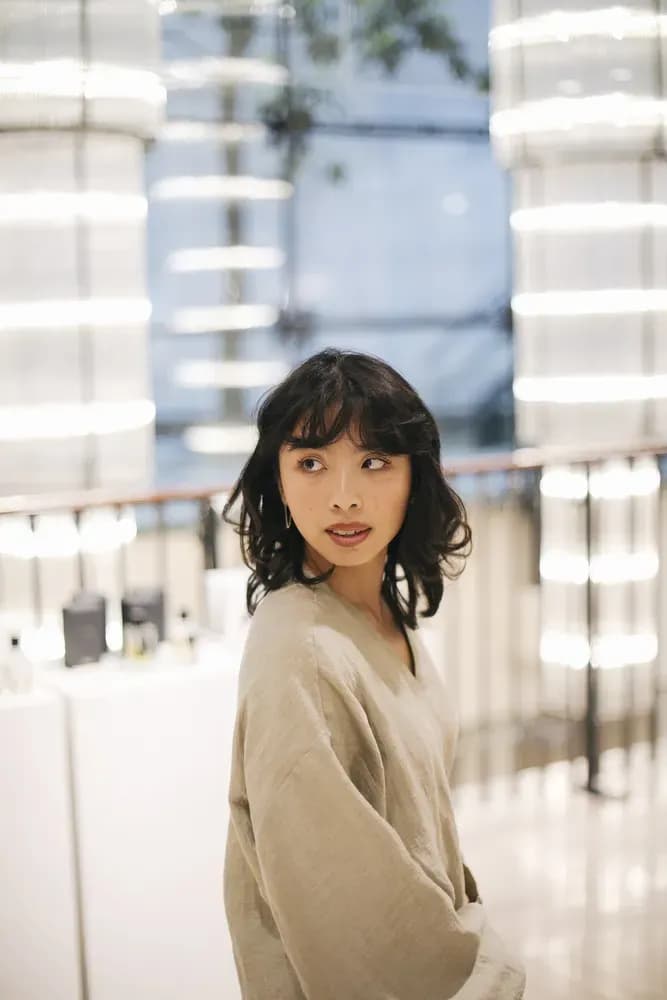 Woman with short wavy dark hair wearing a loose beige top, looking over her shoulder in a softly lit, modern indoor space.