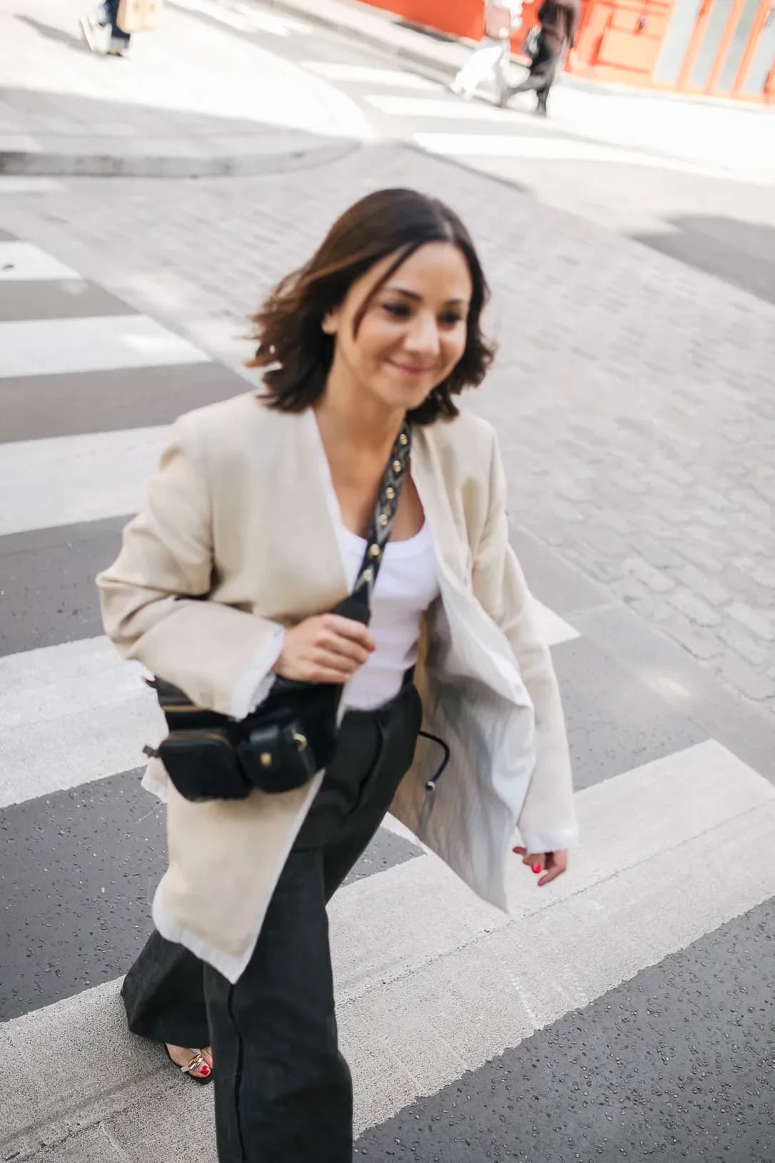 Woman in a beige blazer and relaxed trousers walking across a city street with a camera, showcasing a modern urban style.