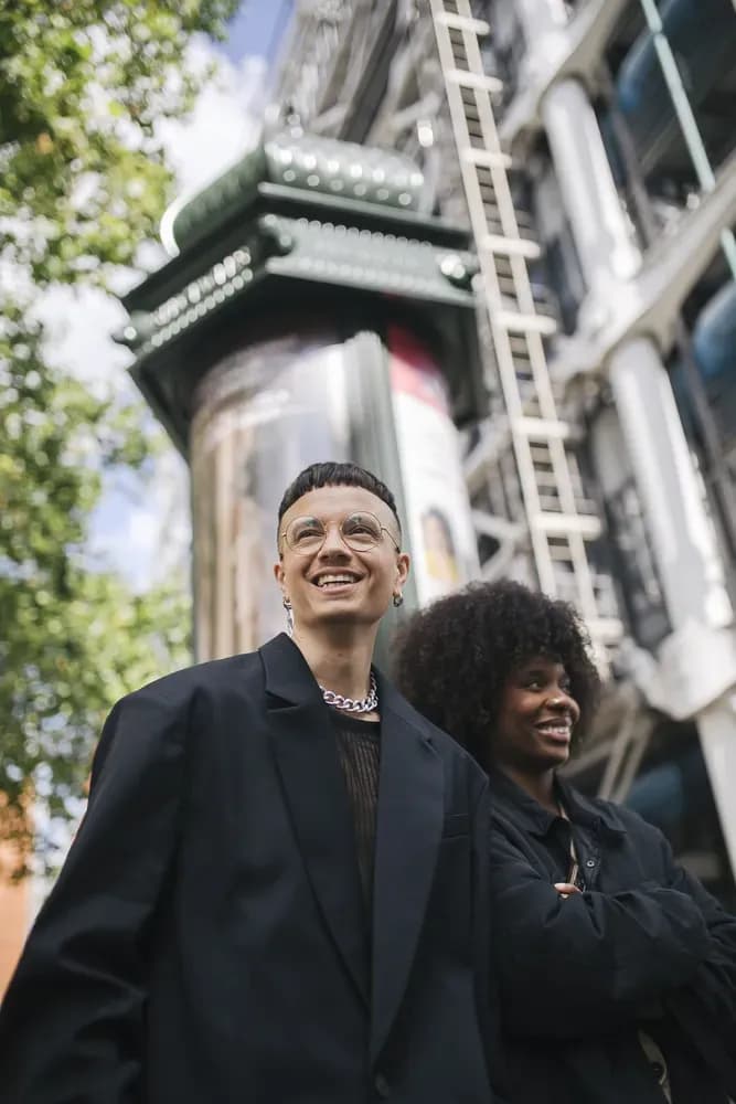 Two people in black outfits smiling outdoors in a city setting, styled in a modern, urban fashion look.