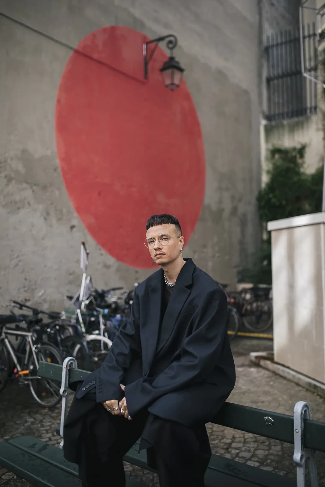 Man in an oversized black suit sitting on a bench in an urban setting with a bold red wall, showcasing a modern and statement street style.