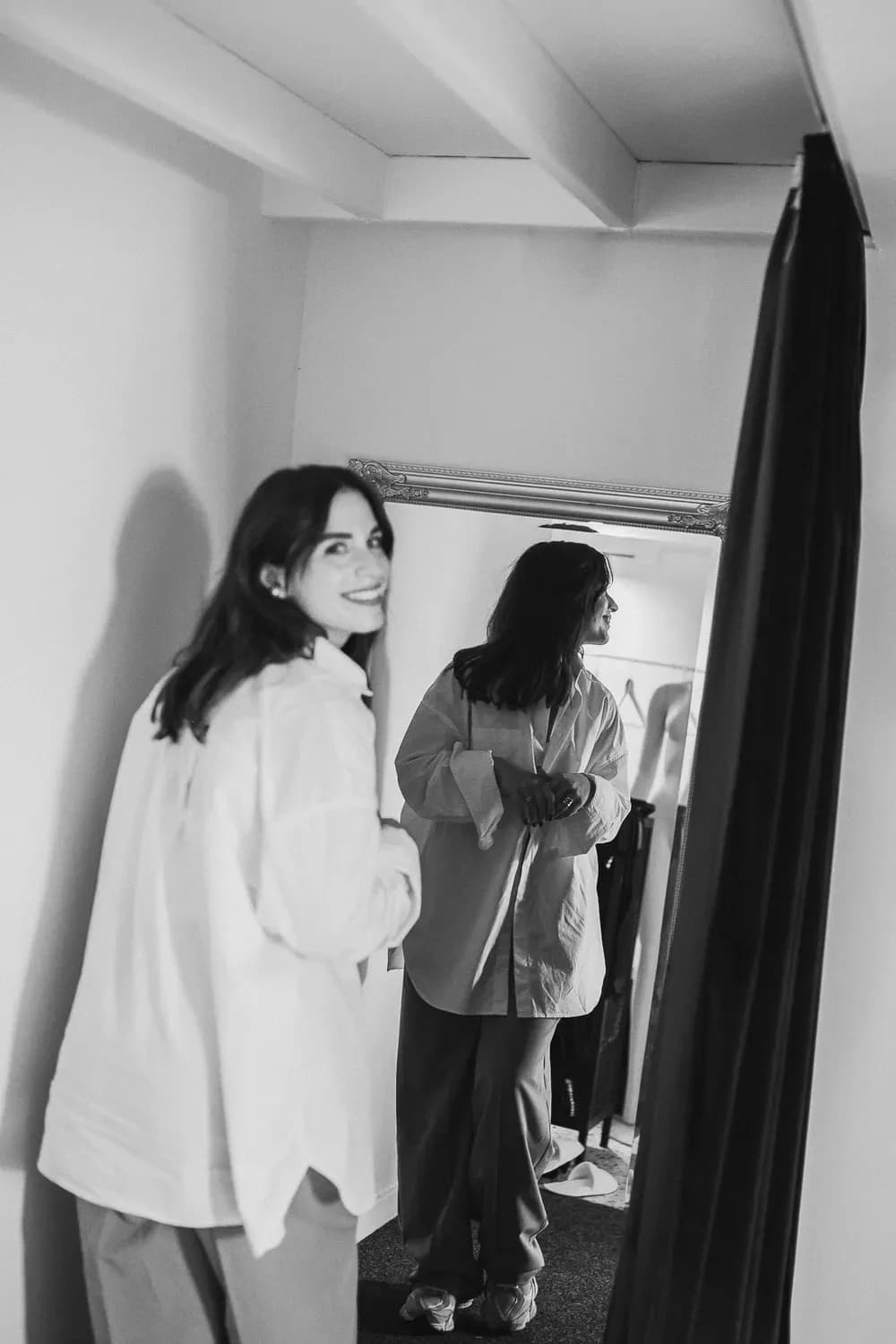 Χαμογελαστή γυναίκα με χαλαρό, φαρδύ πουκάμισο στέκεται σε ένα δοκιμαστήριο, κοιτάζοντας πίσω ενώ ελέγχει το ντύσιμό της σε έναν ολόσωμο καθρέφτη.