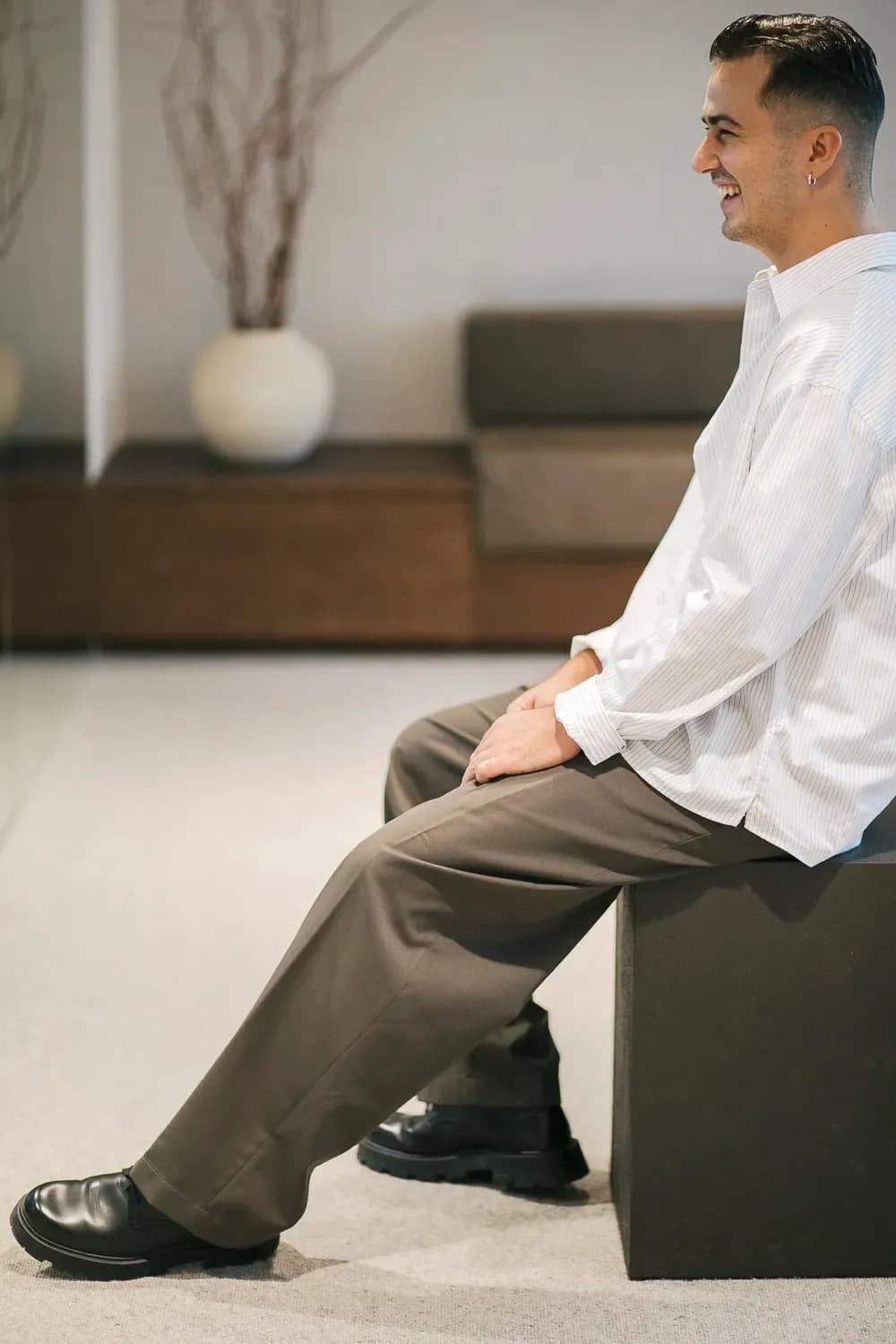 Άνδρας με χαλαρό ριγέ πουκάμισο και ραμμένο παντελόνι κάθεται σε ένα παγκάκι σε εσωτερικό χώρο, χαμογελώντας σε ένα ήρεμο, μινιμαλιστικό περιβάλλον.