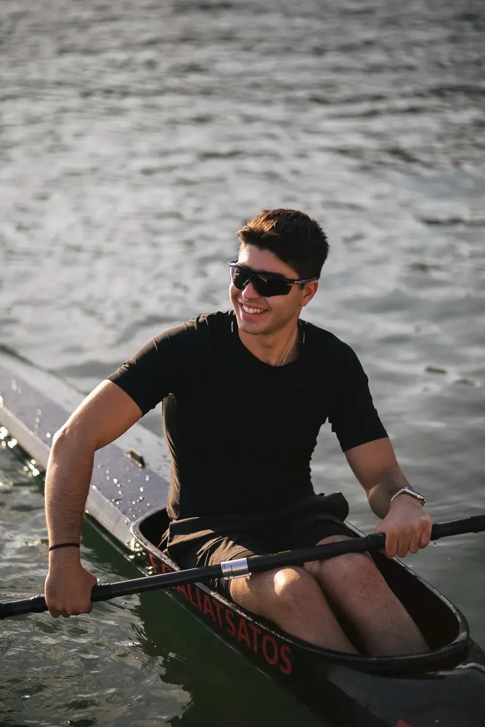 Άνδρας με μαύρο μπλουζάκι και γυαλιά ηλίου κάνει καγιάκ στο νερό, χαμογελώντας σε μια χαλαρή, αθλητική στιγμή σε εξωτερικό χώρο.