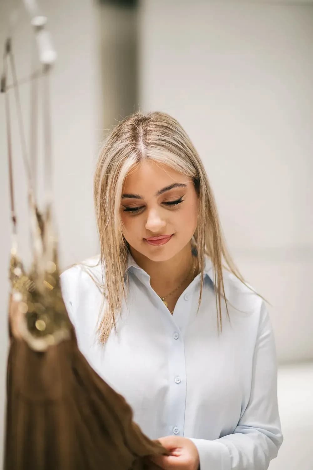 Γυναίκα με ανοιχτό μπλε πουκάμισο εξετάζει ένα ένδυμα σε κρεμάστρα σε ένα απαλά φωτισμένο δοκιμαστήριο, εστιάζοντας στο ύφασμα και τη λεπτομέρεια.