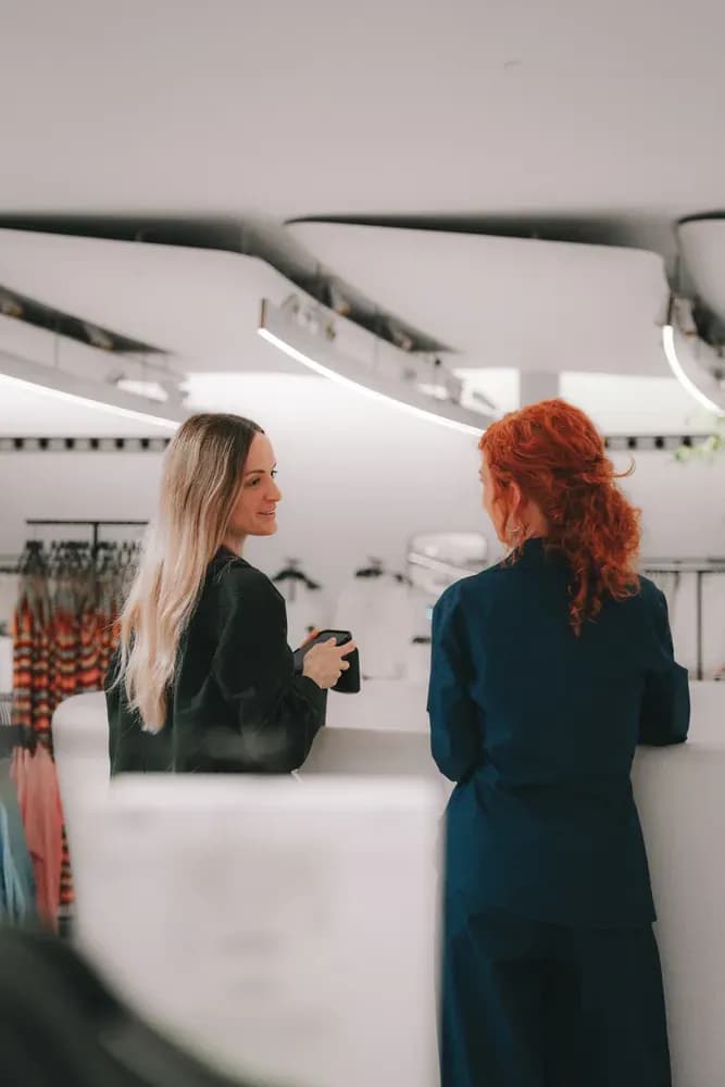 Two women chatting at the counter of a contemporary fashion store, dressed in tailored minimalist outfits.