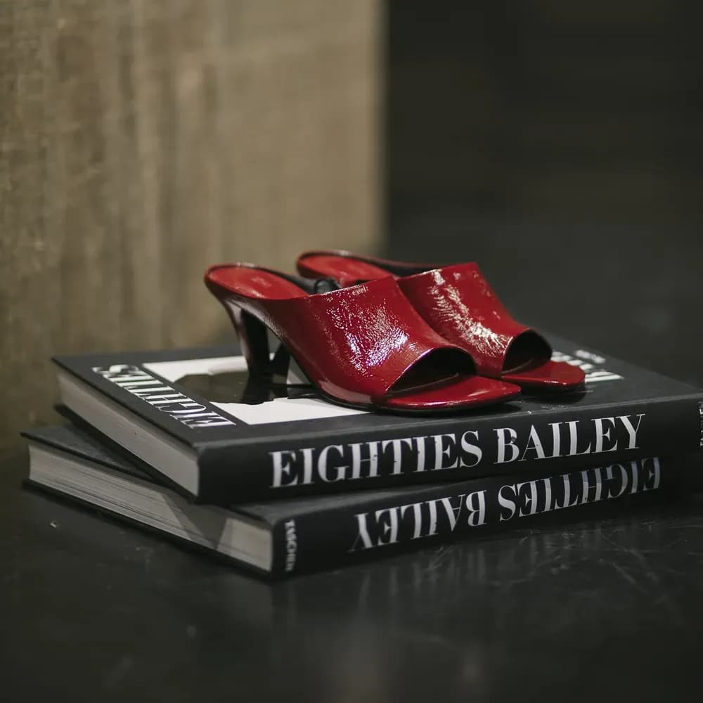 Glossy red leather open-toe heeled mules displayed on stacked fashion books.