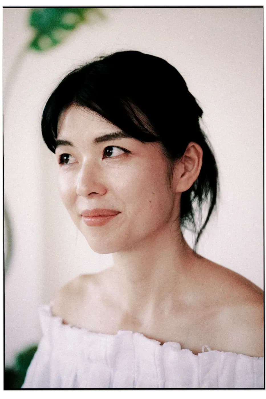 Close-up portrait of a person with hair tied back, wearing an off-shoulder white top, looking to the side in a soft, minimal setting.