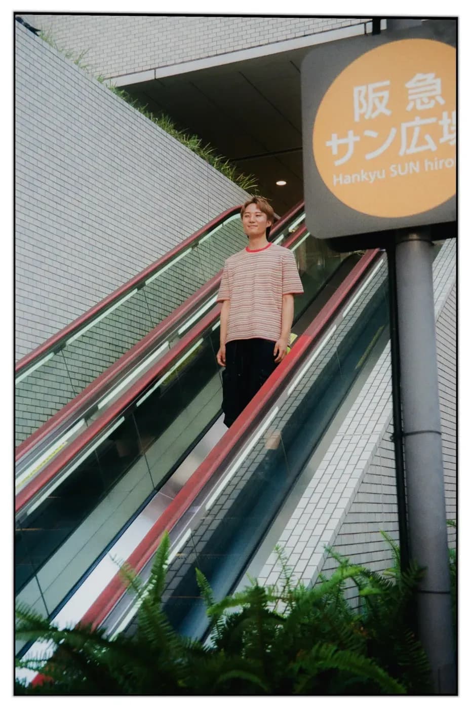 Person riding down an outdoor escalator, wearing a striped t-shirt and dark pants, next to a sign in an urban setting.
