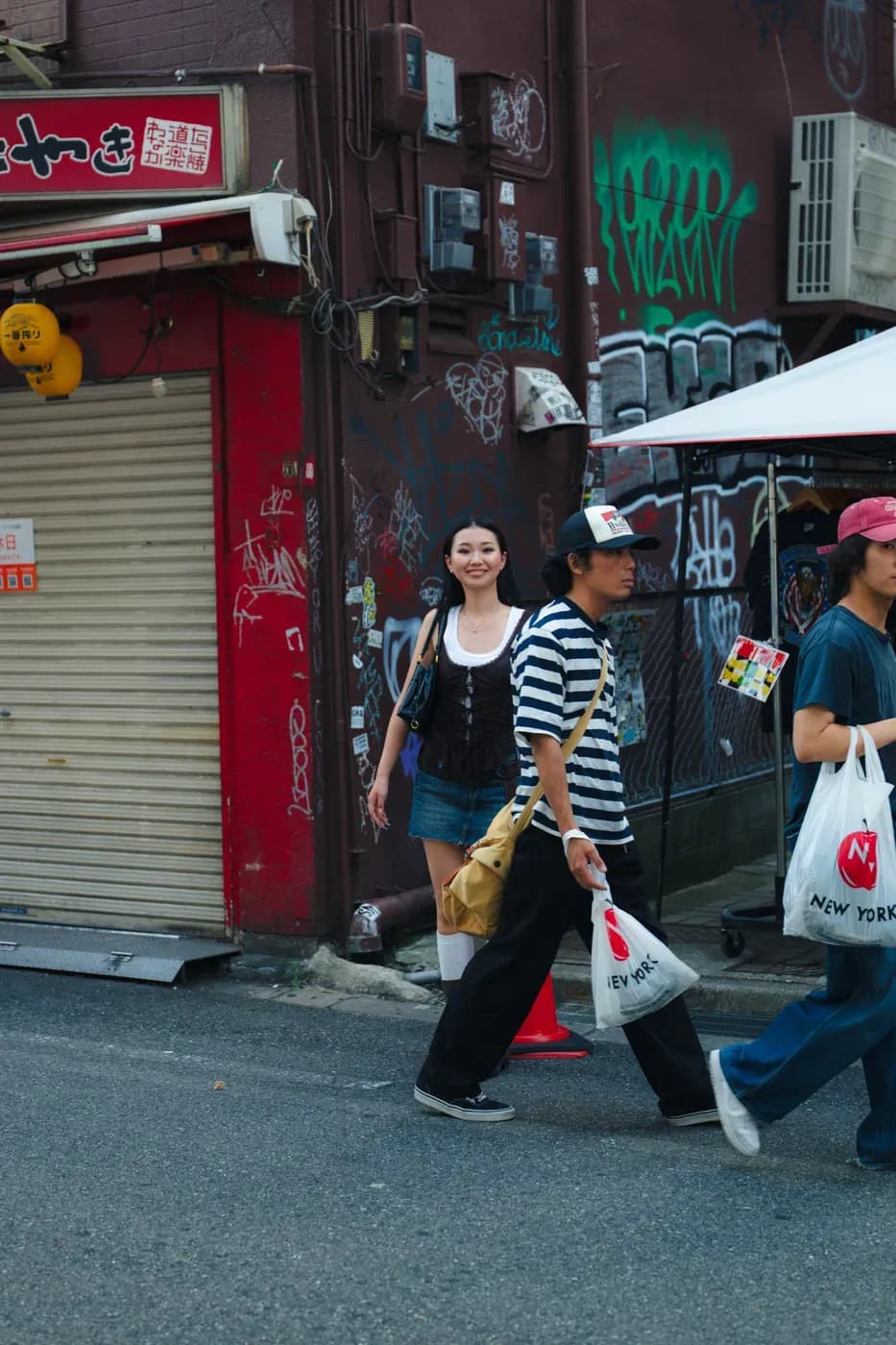 Person walking along a graffiti-covered street, wearing a casual outfit with layered pieces, while others pass by carrying shopping bags in an urban setting.