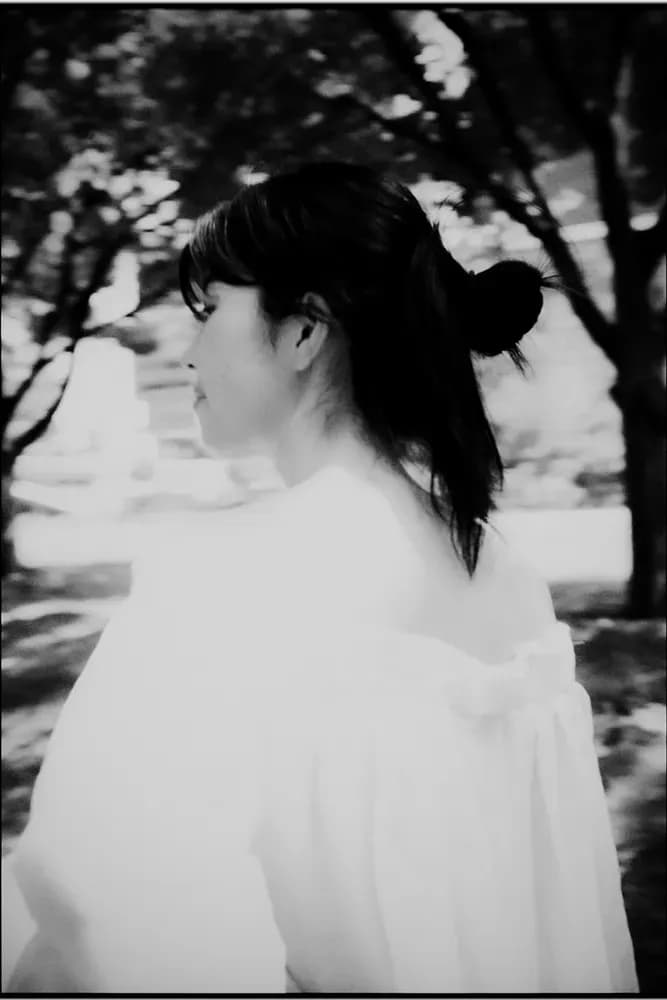 Black-and-white profile of a woman with dark hair in a low bun, wearing a loose white blouse, standing outdoors among softly blurred trees.