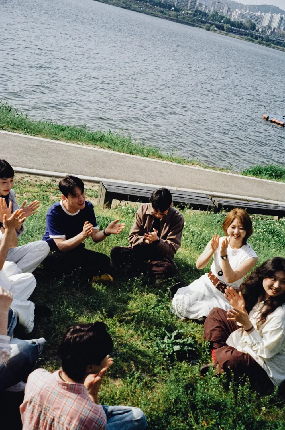 Group of friends sitting on the grass by a riverside, clapping and smiling together in relaxed contemporary casual outfits.