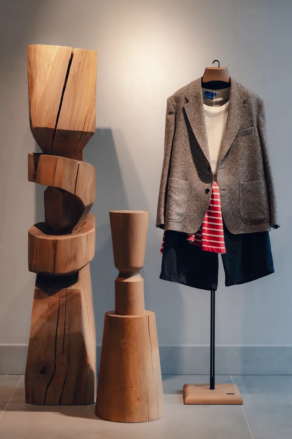 Grey textured blazer layered with casual separates displayed beside sculptural wooden furniture in a modern fashion retail setting.
