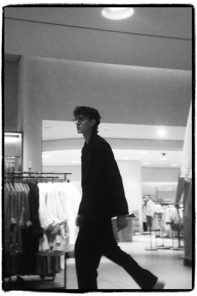 Imagen en blanco y negro de un hombre con ropa oscura caminando por un espacio comercial minimalista con percheros al fondo.