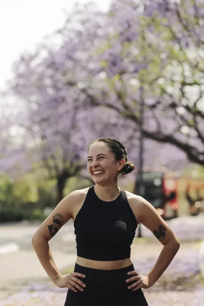 Smiling woman in a black sleeveless top standing outdoors beneath blooming trees, hands on hips in soft natural light