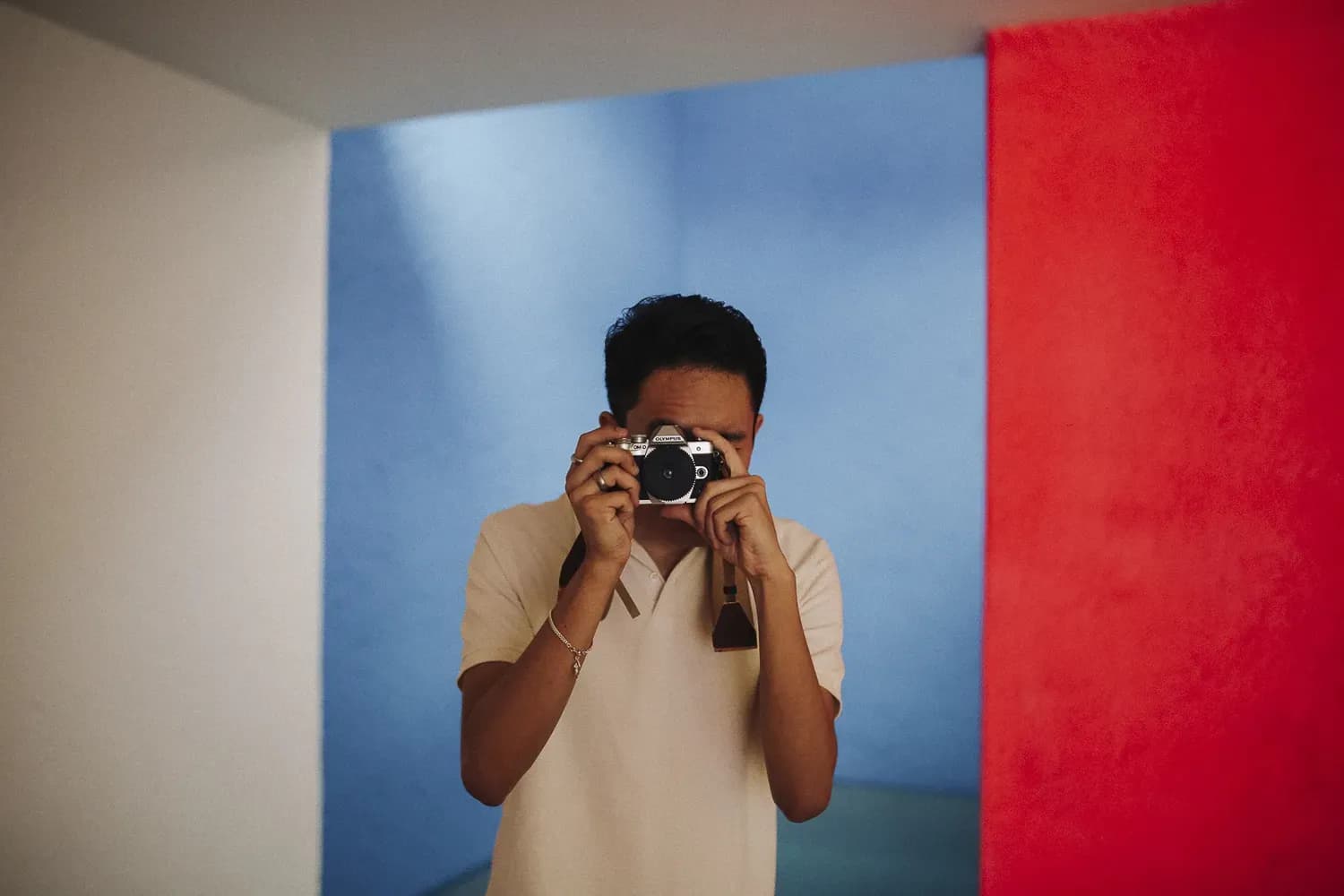 Man in a beige polo holding a camera up to his face, standing against a bold blue and red background.