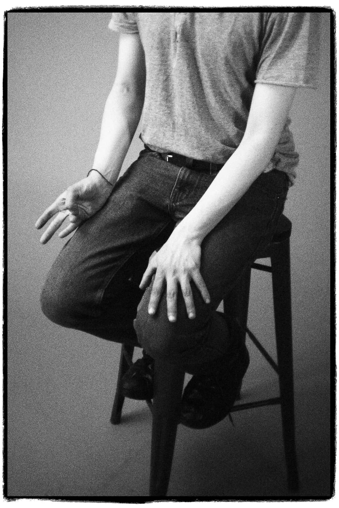 Primer plano en blanco y negro de una persona sentada con una camiseta ajustada y vaqueros oscuros, con las manos relajadas sobre el regazo en un taburete sencillo.