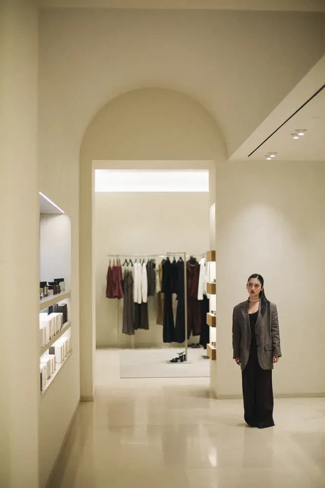 Woman in a blazer standing in a minimalist boutique interior with neutral tones and curated clothing displays.