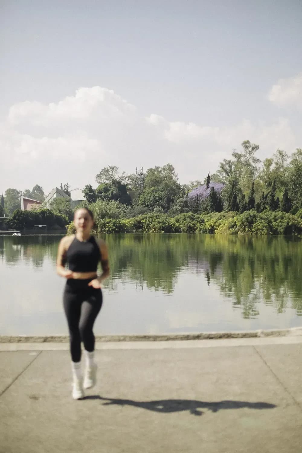 Person running by a lake, wearing black sportswear and light sneakers, in a natural environment with trees and clear sky.