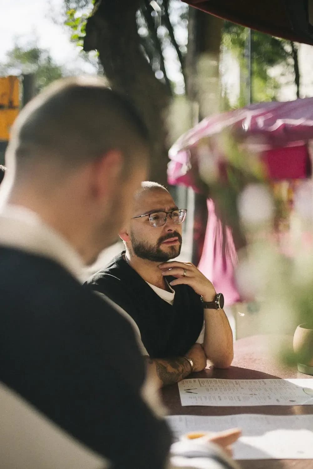 A person with glasses and a trimmed beard is conversing at an outdoor table, wearing a dark t-shirt in an urban environment with natural light.
