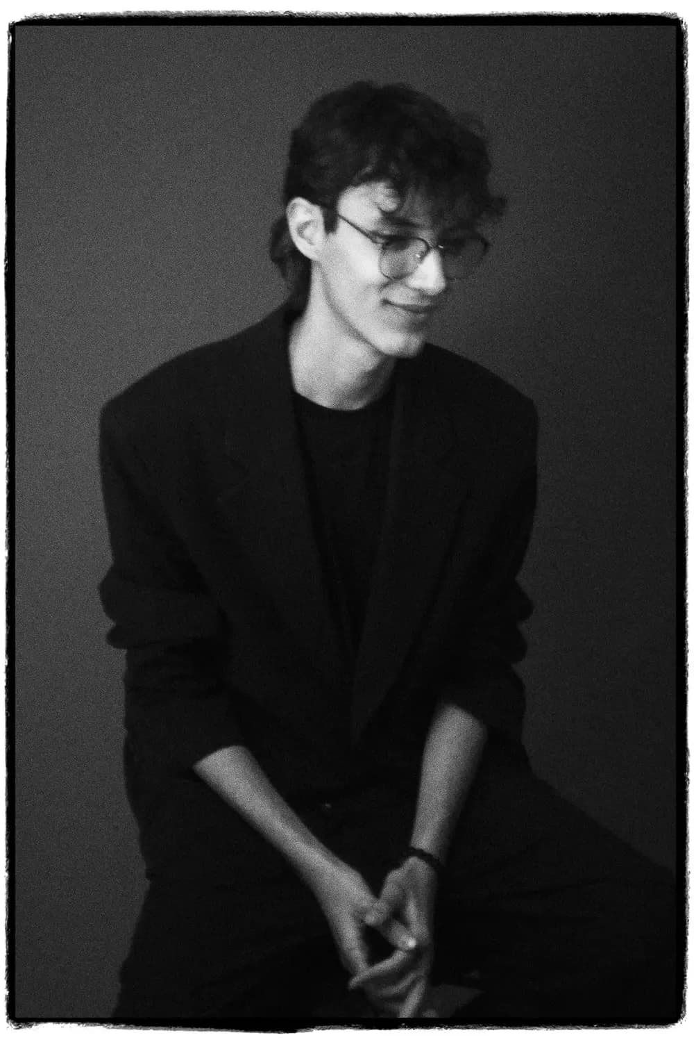 Young person sitting, wearing a black suit with a dark t-shirt and glasses, in a black and white portrait on a neutral background.