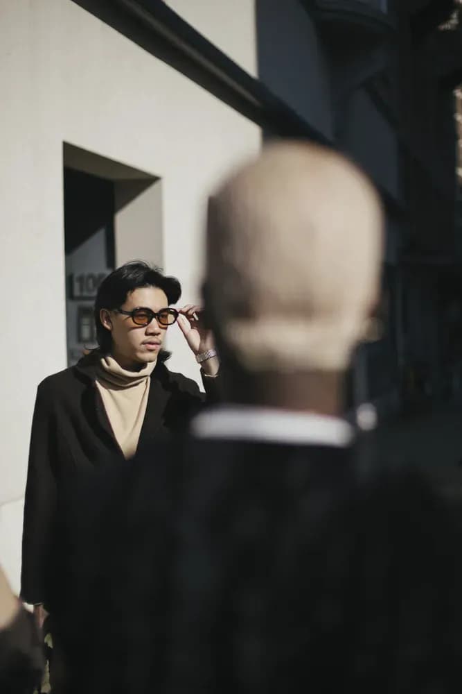 Person outdoors adjusts sunglasses, wears dark coat over high collar in neutral tones, with illuminated urban background.