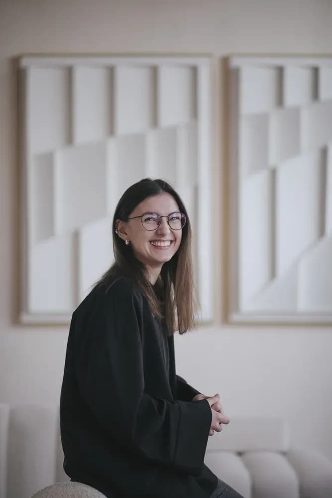Smiling person wearing glasses and a black outfit, seated in a minimalist interior with geometric wall art in the background.