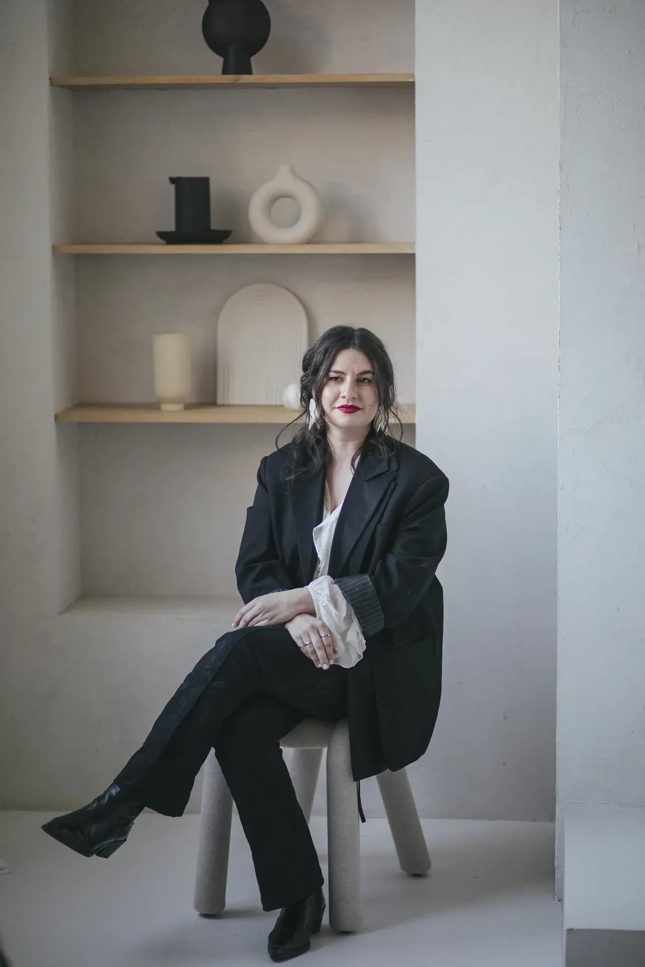 A woman sits on a stool wearing a tailored black suit with a white blouse, posing in a minimalist interior with neutral-toned shelves and decorative objects in the background.
