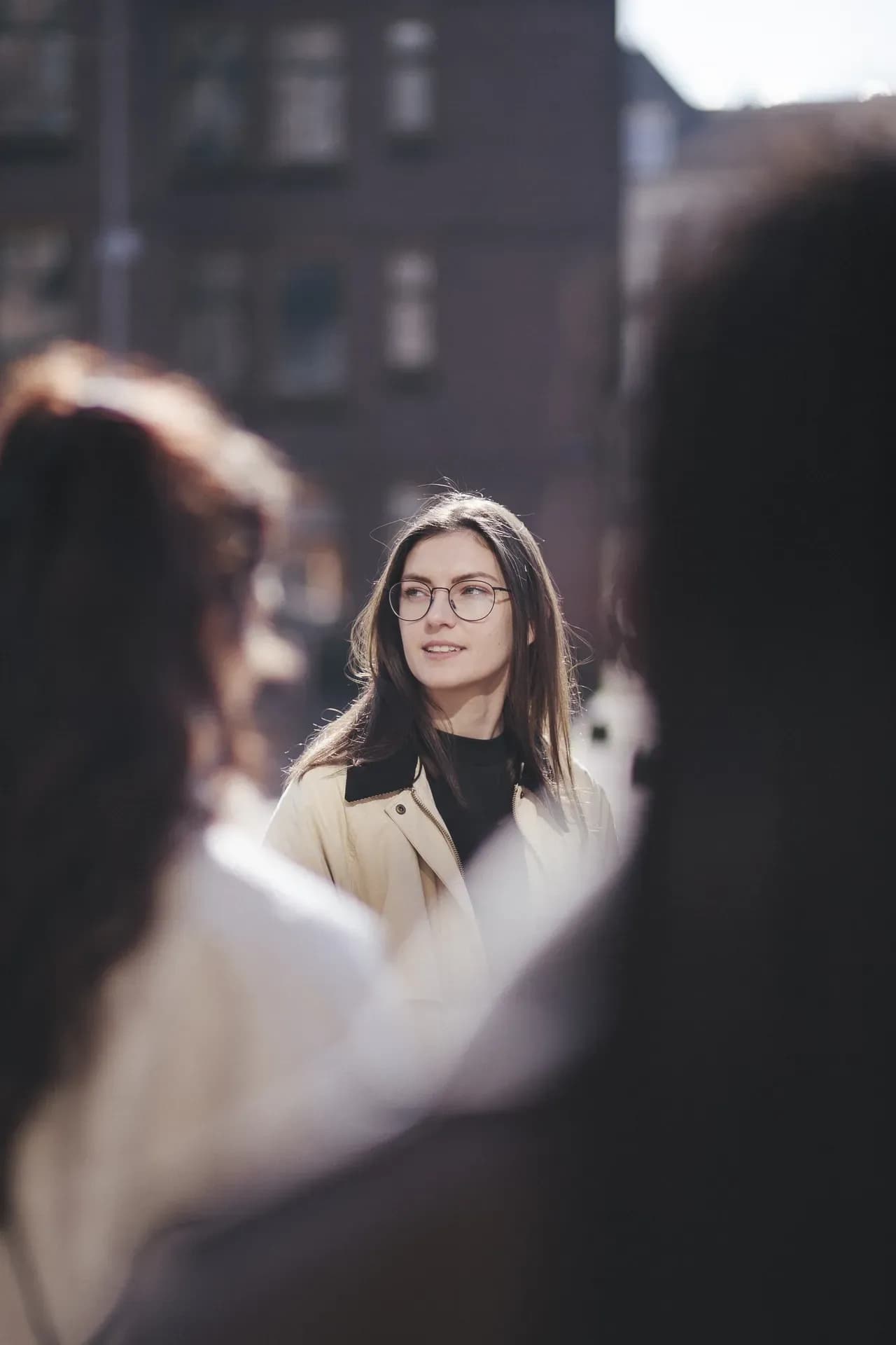 A woman wearing glasses and a light jacket stands outdoors in a city, looking to the side, with blurred figures and buildings in the background.
