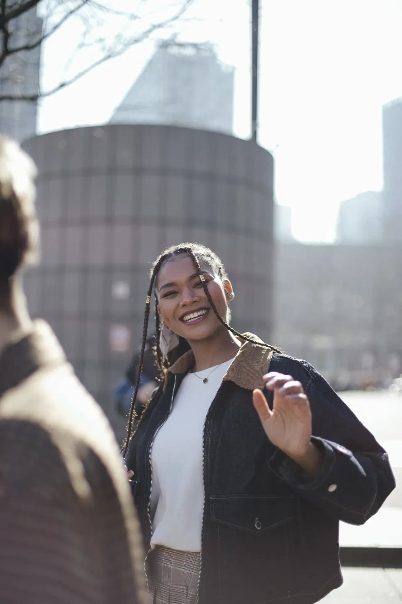 A woman wearing a dark jacket with a contrasting collar and a white top smiles and gestures outdoors in a bright urban setting, with buildings in the background.