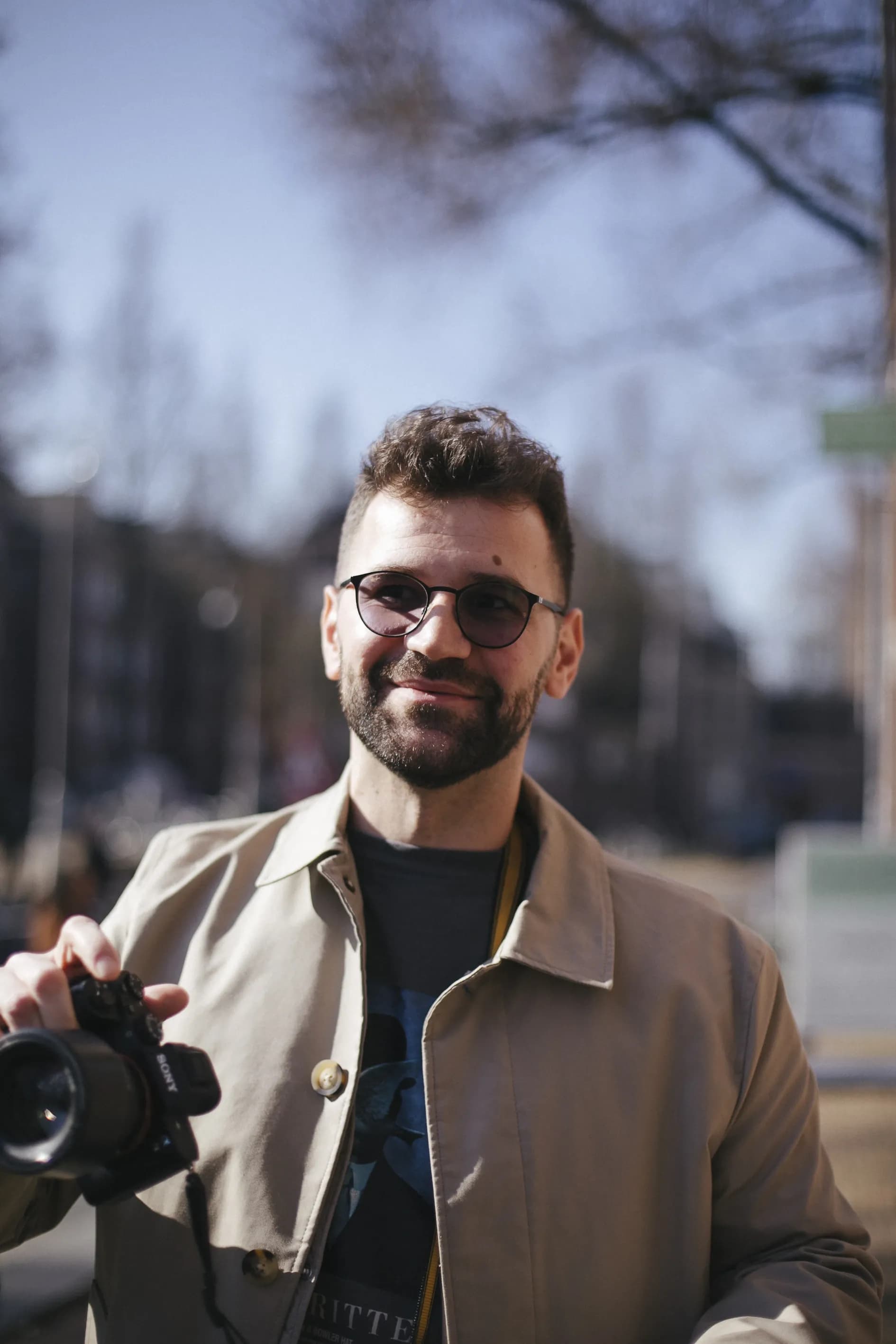 A man wearing sunglasses and a beige jacket smiles while holding a camera in an outdoor urban setting.