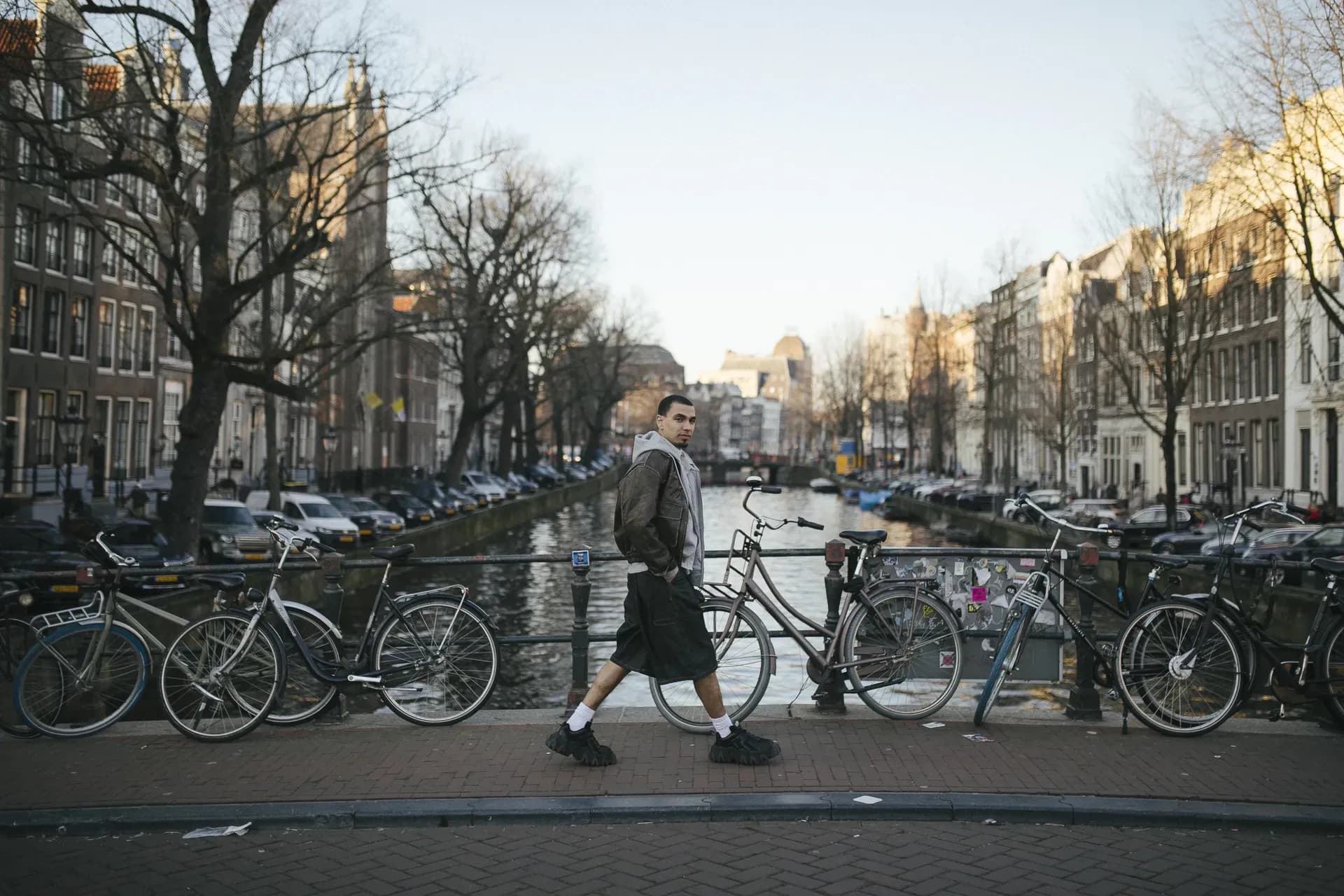 A person walks along a canal-lined city street, wearing a casual, layered outfit that blends utility and comfort, set against an urban backdrop that emphasizes everyday movement and contemporary style.