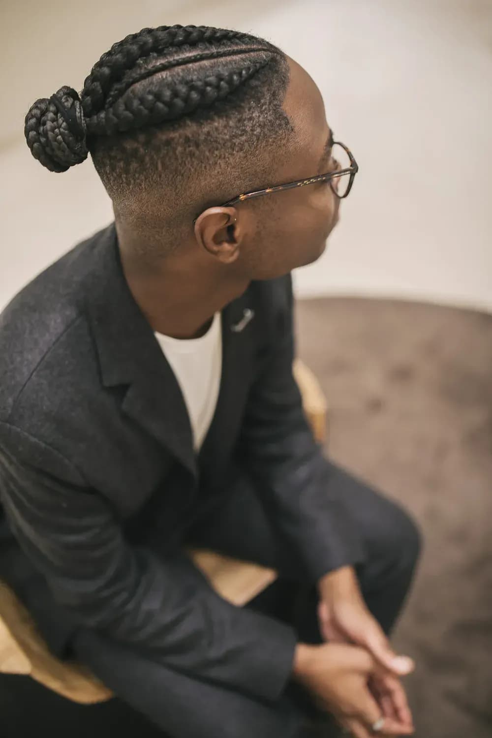 Person with braided hair and glasses wearing a dark suit, seated and looking to the side in a minimalist interior.