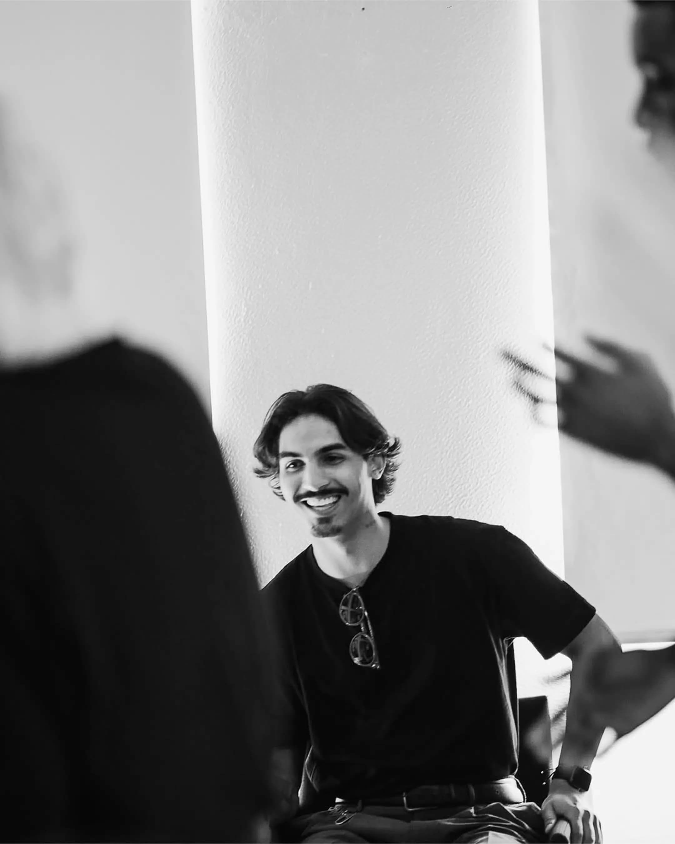 Man smiling and chatting with others in a relaxed indoor setting, captured in a candid black-and-white moment.