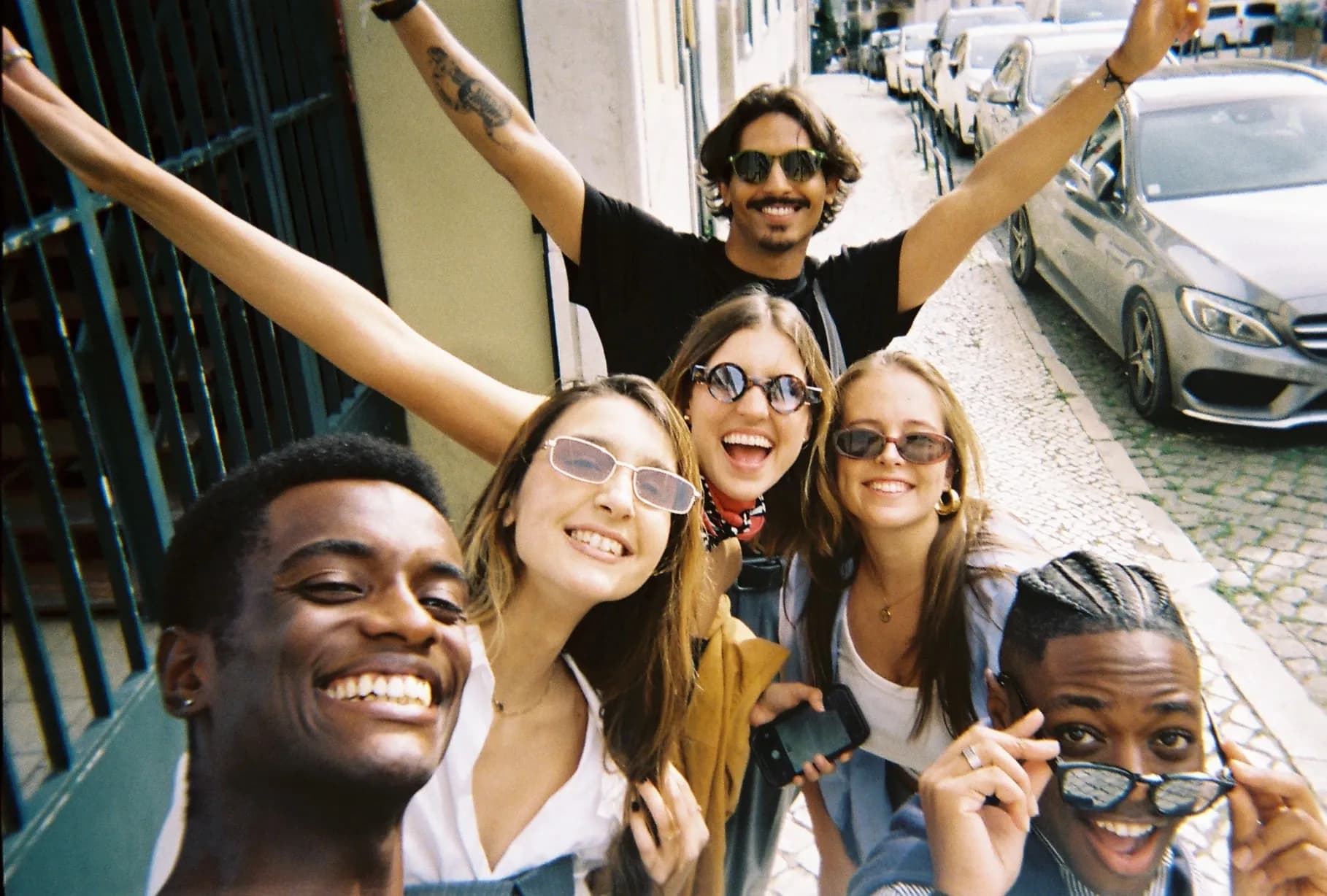 Group of friends smiling and taking a selfie on a sunny city street, wearing casual outfits and sunglasses.