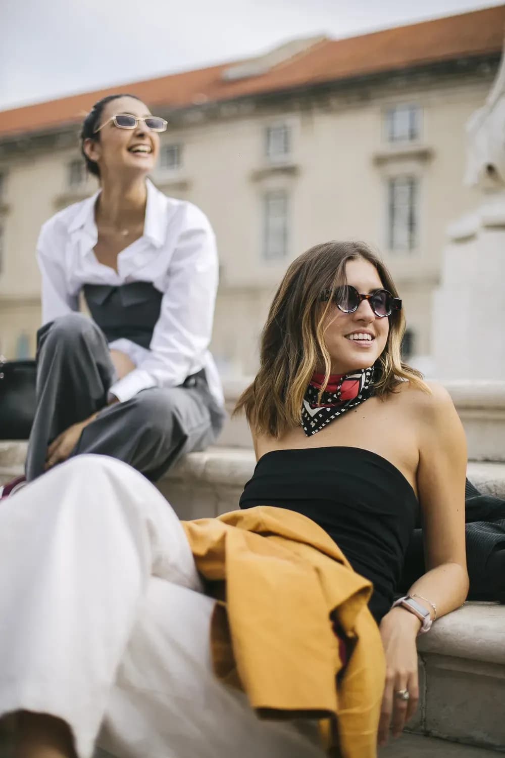 Two women wearing sunglasses sitting outdoors in a city setting, styled in casual-chic outfits with a relaxed, fashionable vibe.